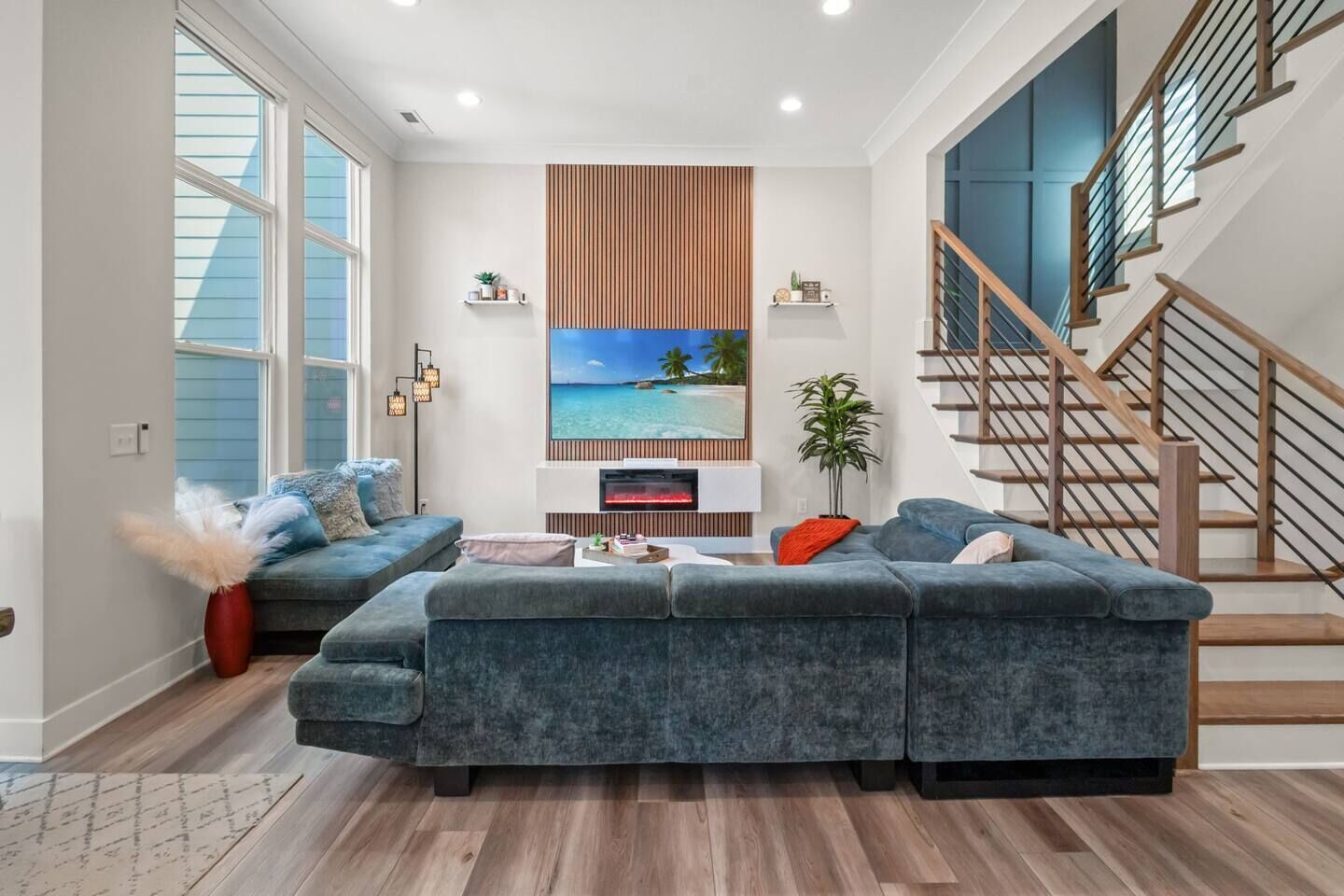A modern living room with a sectional sofa, a flat-screen TV mounted on a wooden panel, and a staircase with wooden railings.