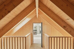 Ein Dachzimmer aus Holz mit schrägen Wänden, einem mittleren Durchgang zu einem angrenzenden Raum und einer Holzvertäfelung an den Wänden.