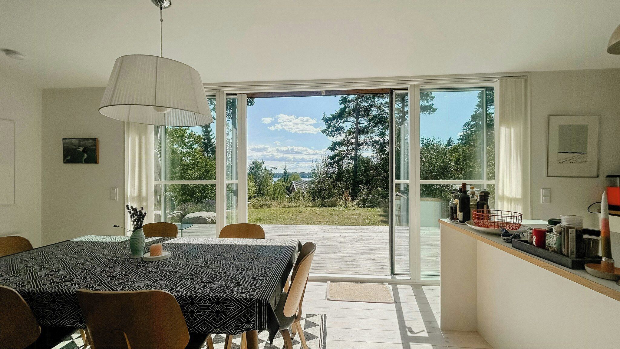 Comedor con mesa y sillas, una lámpara colgante y vista a un jardín mediante puertas de vidrio.