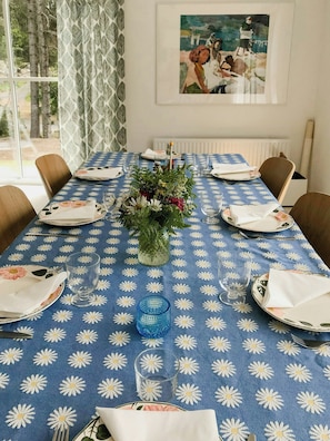 Ein Esstisch ist für sechs Personen gedeckt, mit einer blauen Tischdecke mit Blumenmuster, einer Blumenvase und einem Bild an der Wand.