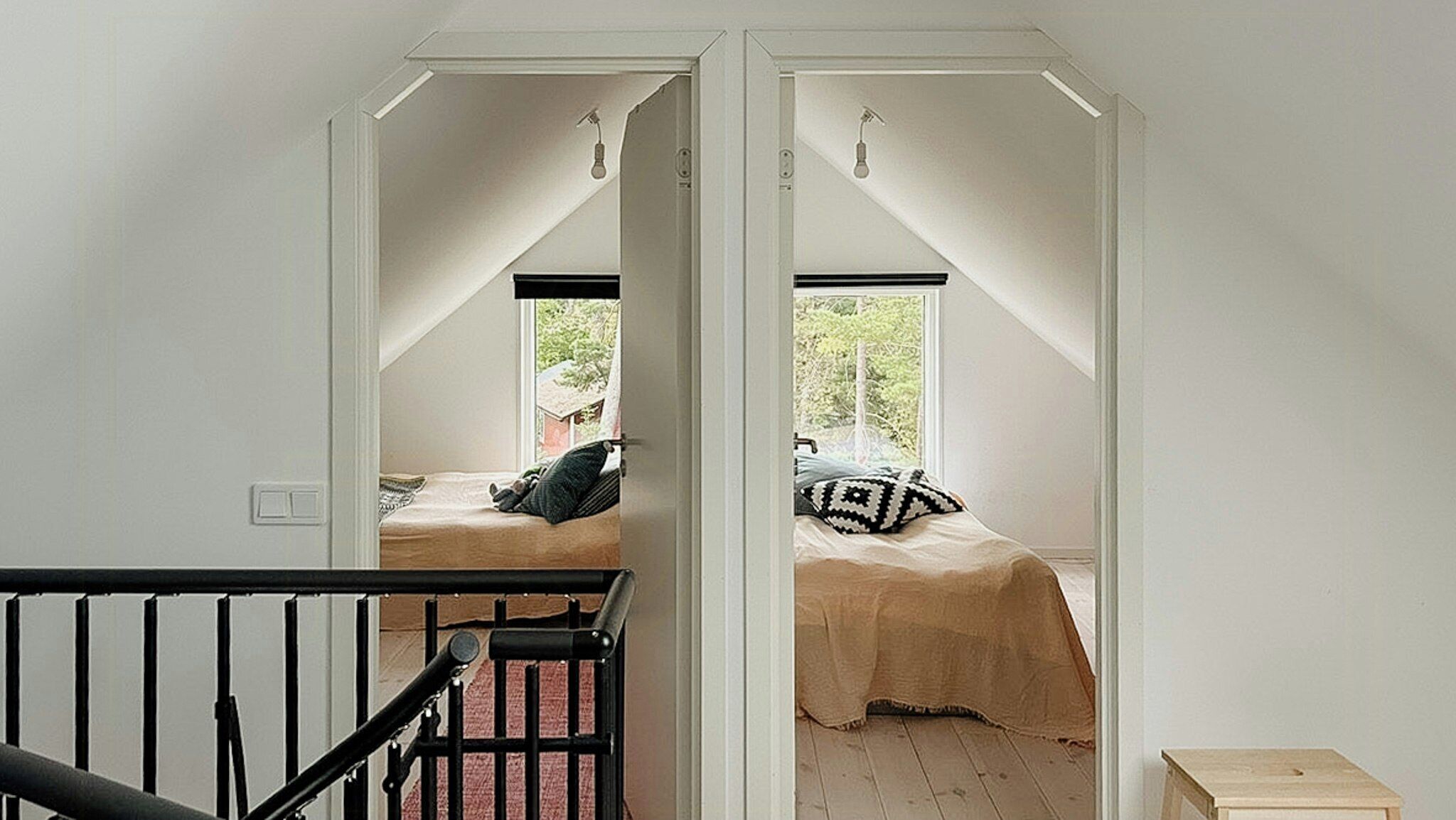Ein Schlafzimmer mit schrägen Wänden, einem Bett mit einem gemusterten Kissen und einem Fenster mit Blick auf Grünflächen.