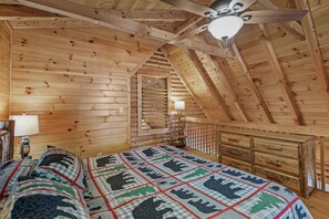Ein Schlafzimmer einer Holzhütte mit schrägem Dach, einem Bett mit einem gemusterten Quilz, einem Nachttisch, einer Lampe und einer Holzkommode.