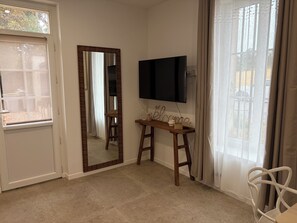 A room with a large mirror, a television mounted on the wall, a small wooden table with decorative items, and a white chair.