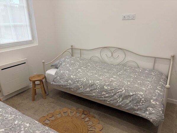 A single bed with a patterned bedspread, a small stool, a radiator, and a power outlet on the wall.