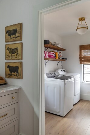 A laundry area with a washing machine and dryer, a wooden shelf with towels and cleaning supplies, and three cow paintings on the wall. - Hotel Bebe Main House - Sleeps 6 (Round Top)