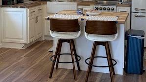 A modern kitchen with white cabinetry, wooden countertops, two bar stools, hanging pendant lights, and a stainless steel stove.