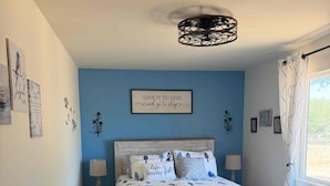 A bedroom with a bed, a nightstand, a lamp, a wall with a framed quote, and a window with curtains.