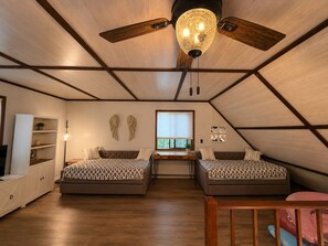 Ein Zimmer mit geneigter Decke, zwei Betten mit gemusterten Bettbezügen, einem Schreibtisch, einem Bücherregal und einem Deckenventilator.