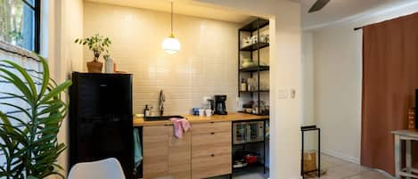 A compact kitchen with a refrigerator, sink, and a coffee maker. There is a dining area with a table and chairs, and a ceiling fan above.