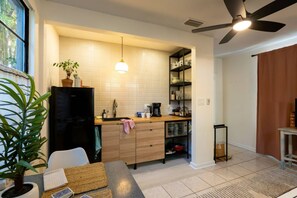 Eine kompakte Küche mit Kühlschrank, Spüle und Kaffeemaschine. Es gibt einen Essbereich mit Tisch und Stühlen, darüber ein Deckenventilator.
