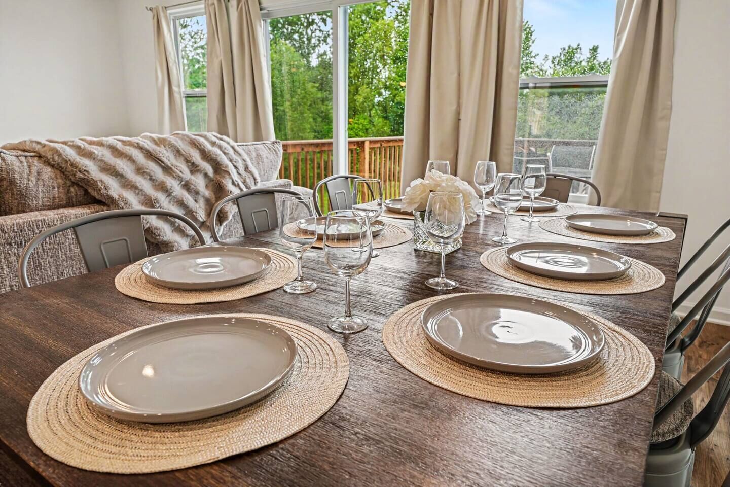 A dining table set with plates, glasses, and a centerpiece, with a view of greenery outside.