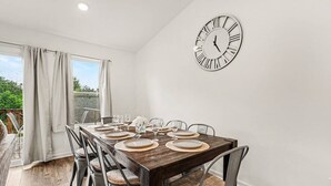 Comedor con mesa de madera dispuesta para cuatro comensales, sillas con asientos de mimbre, un reloj en la pared y vistas a través de una ventana con cortinas.