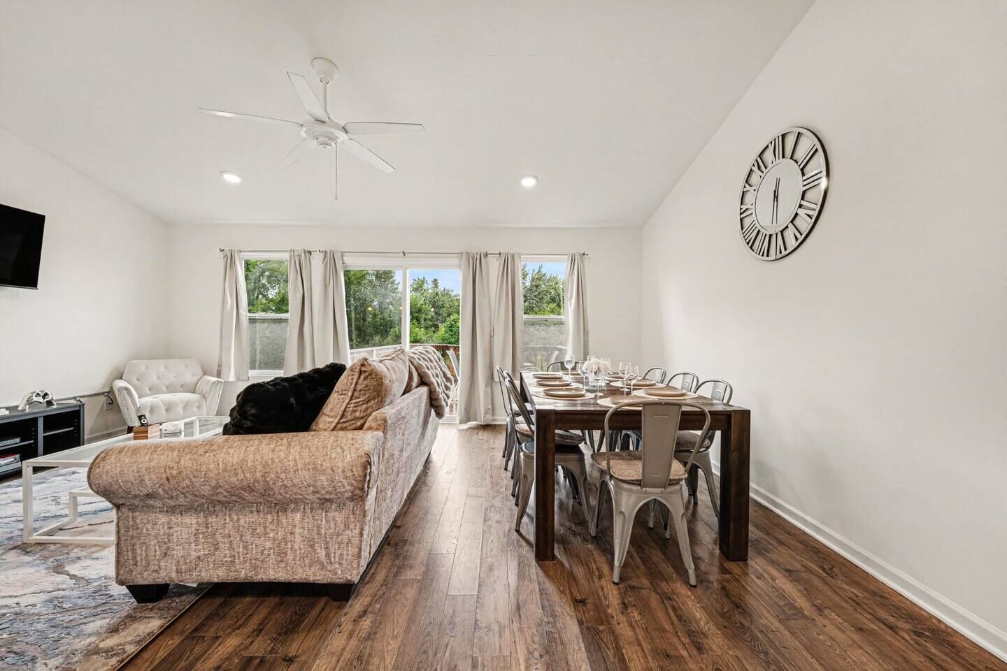 A spacious living room with a sofa, a dining table set for six, and a large wall clock.