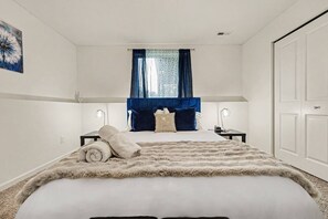 A bedroom with a large bed, two bedside tables, a window with blue curtains, and a door.
