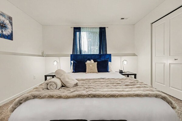 A bedroom with a large bed, two bedside tables, a window with blue curtains, and a door.