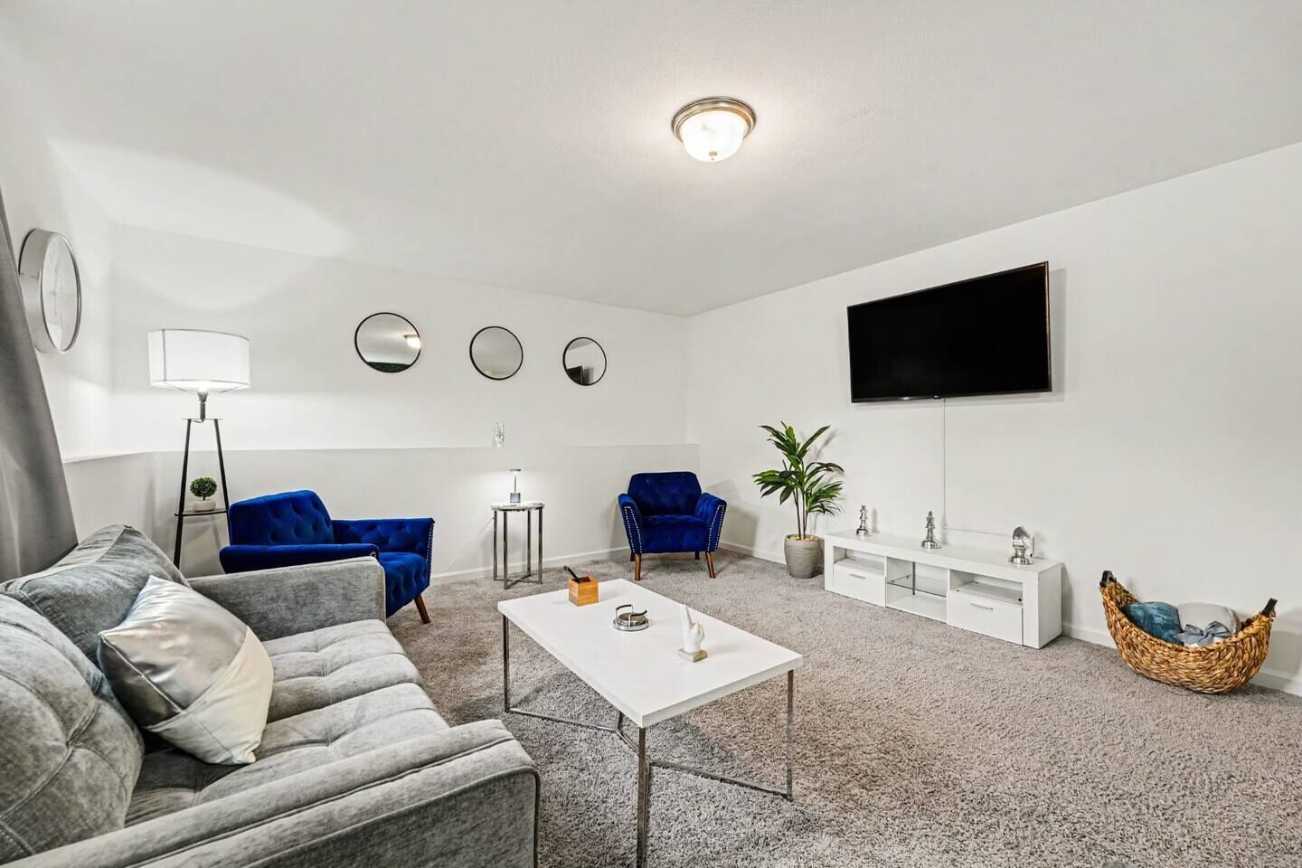 A modern living room with a grey sofa, blue armchairs, a white coffee table, and a flat-screen TV mounted on the wall.