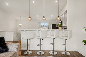 A modern kitchen with white bar stools, pendant lighting, and a view to the outside.