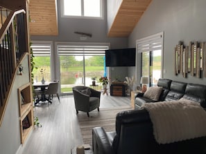 A modern living room with a black leather sofa, a wooden staircase, and a large window overlooking a garden.