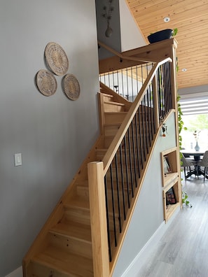 A wooden staircase with black railings, a wall with decorative woven items, and a view of a dining area with chairs and a table.