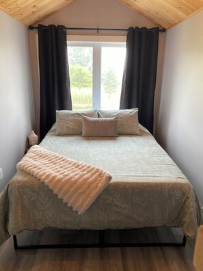 A bedroom with a bed, dark curtains, a window, and a view of greenery outside.