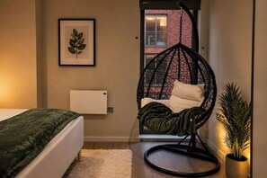 Quarto com cama, cadeira suspensa, radiador e uma impressão botânica emoldurada na parede.