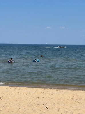 A beach with calm waters, a sandy shore, and a sailboat in the distance.