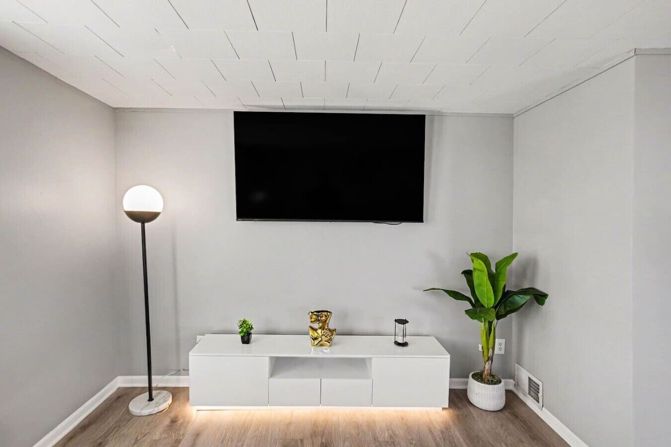 A modern living room with a white entertainment unit, a flat-screen TV mounted on the wall, a floor lamp, and a potted plant.