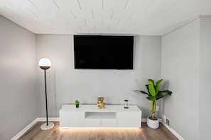 A modern living room with a white entertainment unit, a flat-screen TV mounted on the wall, a floor lamp, and a potted plant.