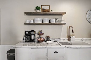 A kitchen with a coffee maker, a sink with a faucet, and a shelf with plates and cups.