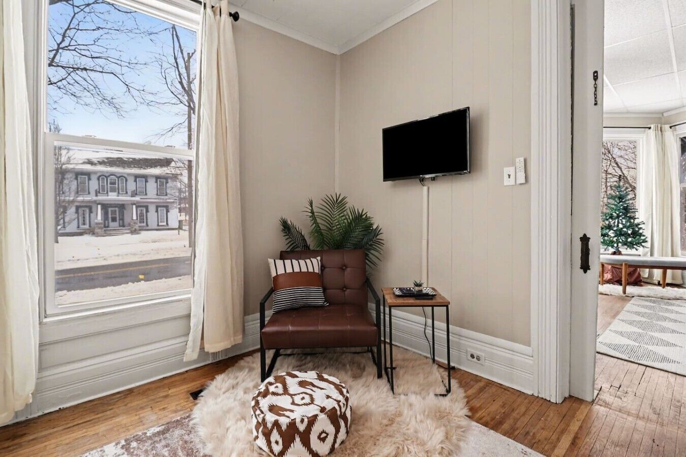A room with a brown leather sofa, a television on the wall, a small wooden side table, and a large window with curtains.