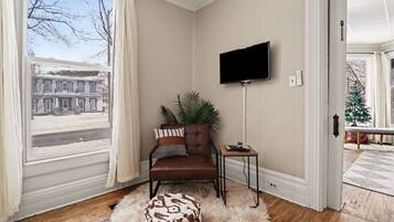 A room with a brown leather sofa, a television on the wall, a small wooden side table, and a large window with curtains.