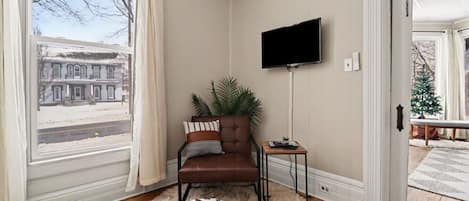 Une pièce avec un canapé en cuir marron, un téléviseur fixé au mur, une petite table d’appoint en bois et une grande fenêtre avec des rideaux.
