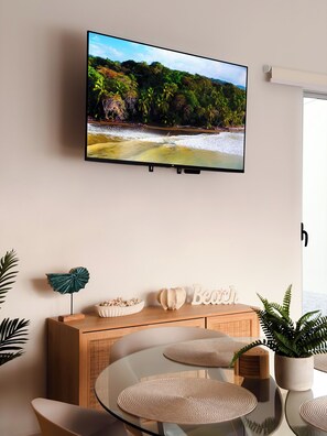 Ein modernes Wohnzimmer mit einem wandmontierten Flachbildfernseher, einem Holztischchen mit Dekoration, einem Glastisch und einem Topfpflanzen.
