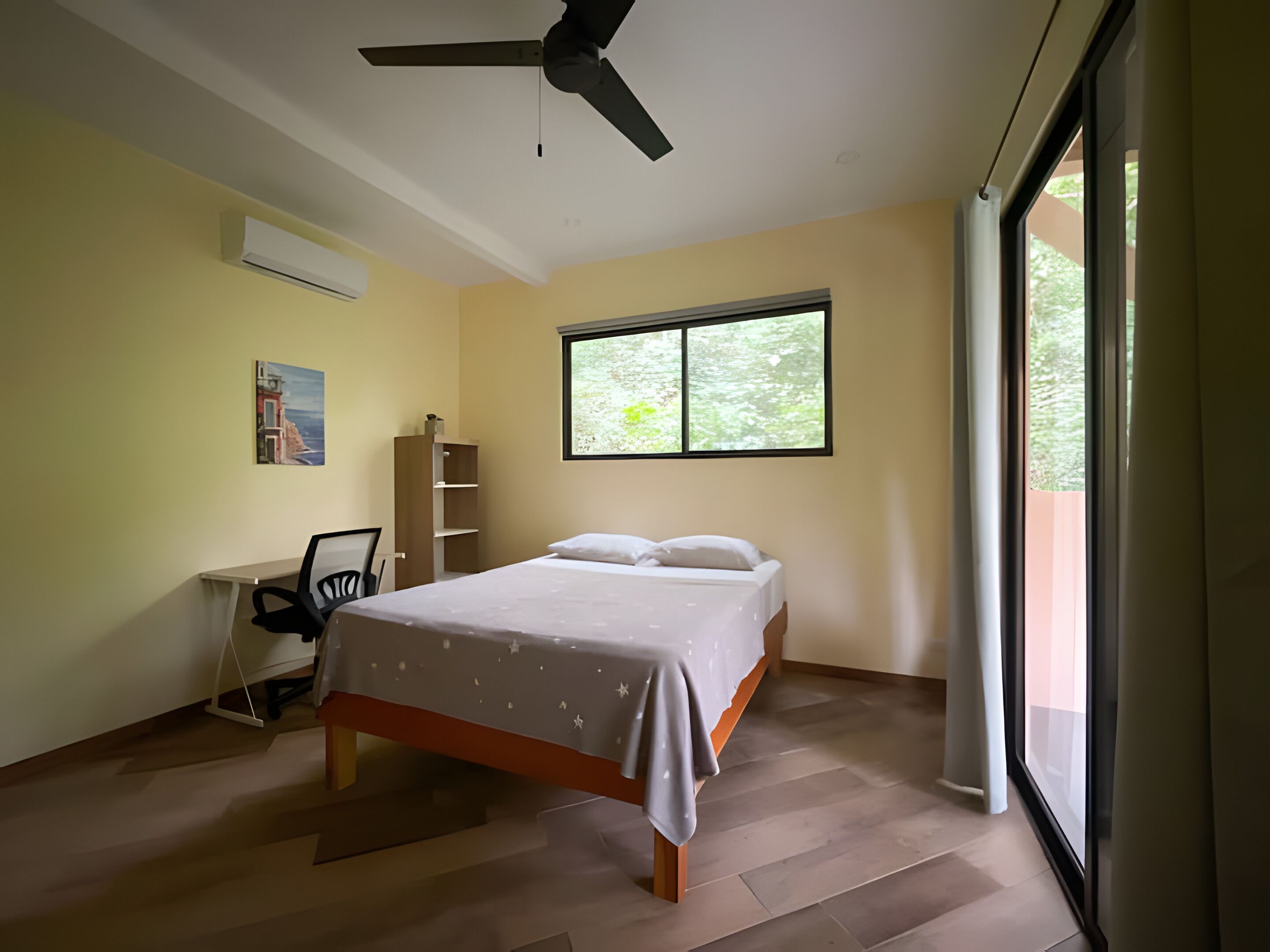 Un dormitorio con cama, escritorio, silla, ventilador de techo y ventana con vistas a vegetación.