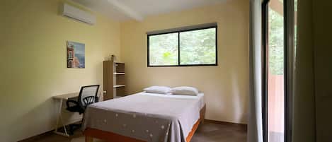 Un dormitorio con cama, escritorio, silla, ventilador de techo y ventana con vistas a vegetación.