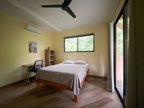 A bedroom with a bed, a desk, a chair, a ceiling fan, and a window with a view of greenery.