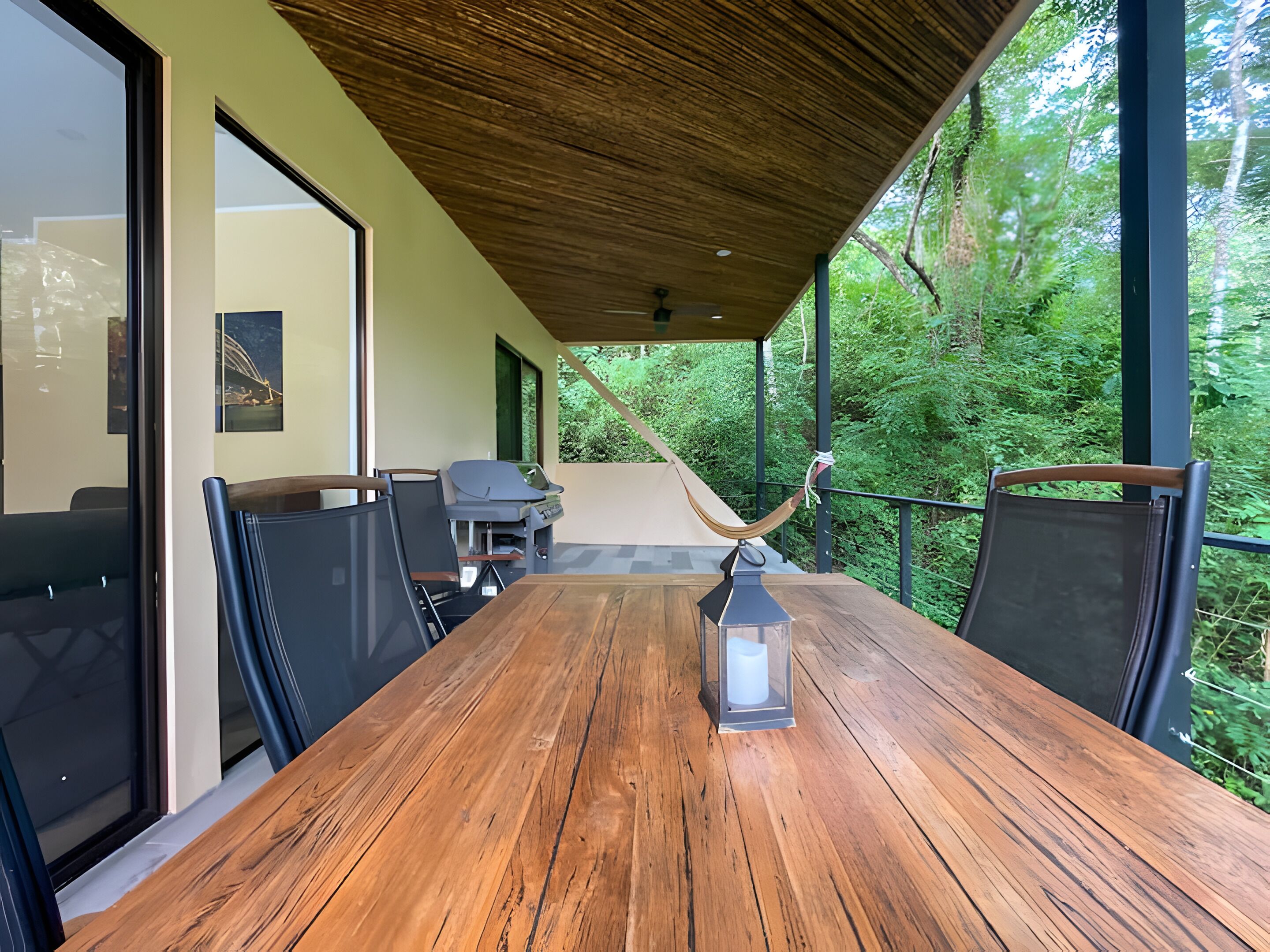 Mesa de madera con sillas, una hamaca y un farol en un espacio exterior cubierto con vista a la vegetación.