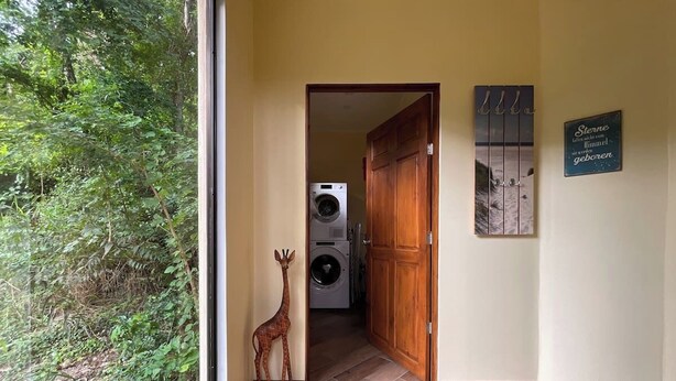 Un couloir avec une porte en bois donnant sur une buanderie, une décoration représentant une girafe et un mur orné d’une phrase en allemand.