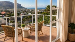 A balcony with wicker chairs and a table, overlooking a coastal town and hills. - Sky Nova Villa False Bay Mountain & Ocean Views (Cape Town)