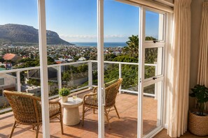 A balcony with wicker chairs and a table, overlooking a coastal town and hills. - Sky Nova Villa False Bay Mountain & Ocean Views (Cape Town)