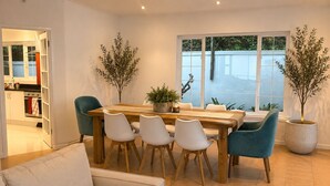 A dining area with a wooden table and white chairs, potted plants, and a view through large windows. - Sky Nova Villa False Bay Mountain & Ocean Views (Cape Town)