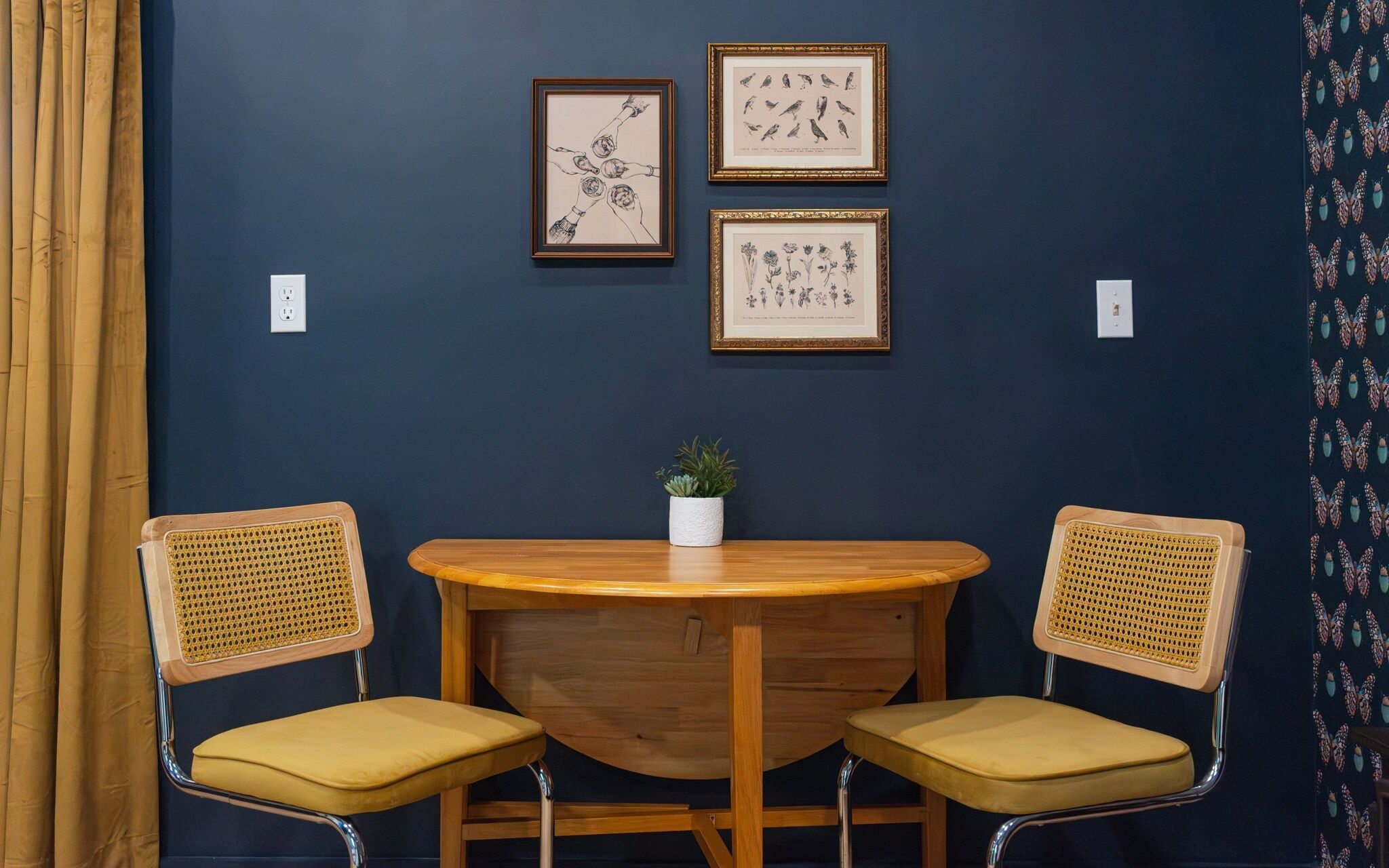 Una habitación con una pared azul oscuro, dos sillas de madera con cojines amarillos, una mesa redonda de madera y tres ilustraciones botánicas enmarcadas.