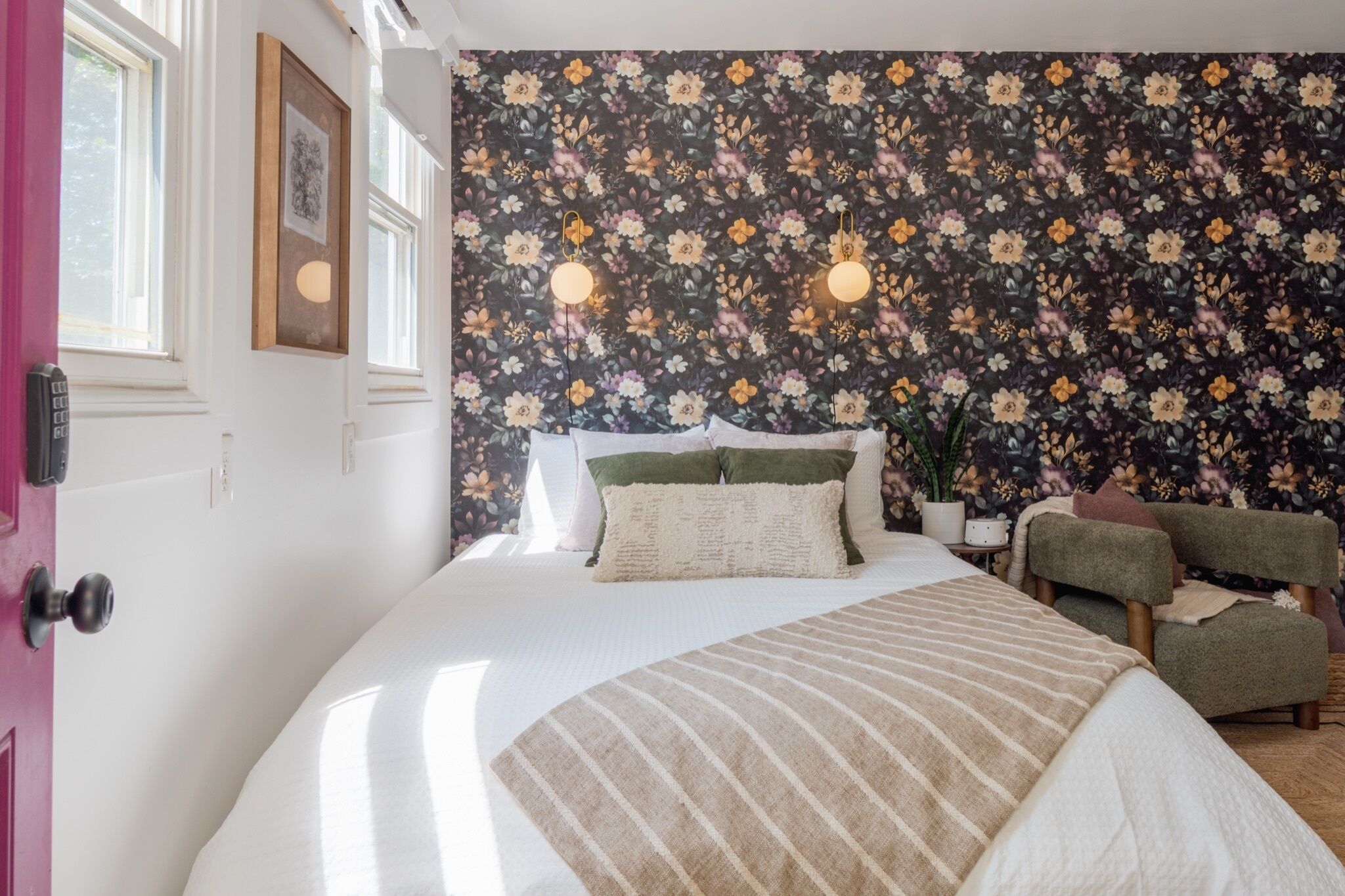 A bedroom with a floral wallpaper, a bed with a striped blanket, a green armchair, and a bedside table with a lamp.