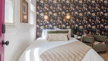 A bedroom with a floral wallpaper, a bed with a striped blanket, a green armchair, and a bedside table with a lamp.