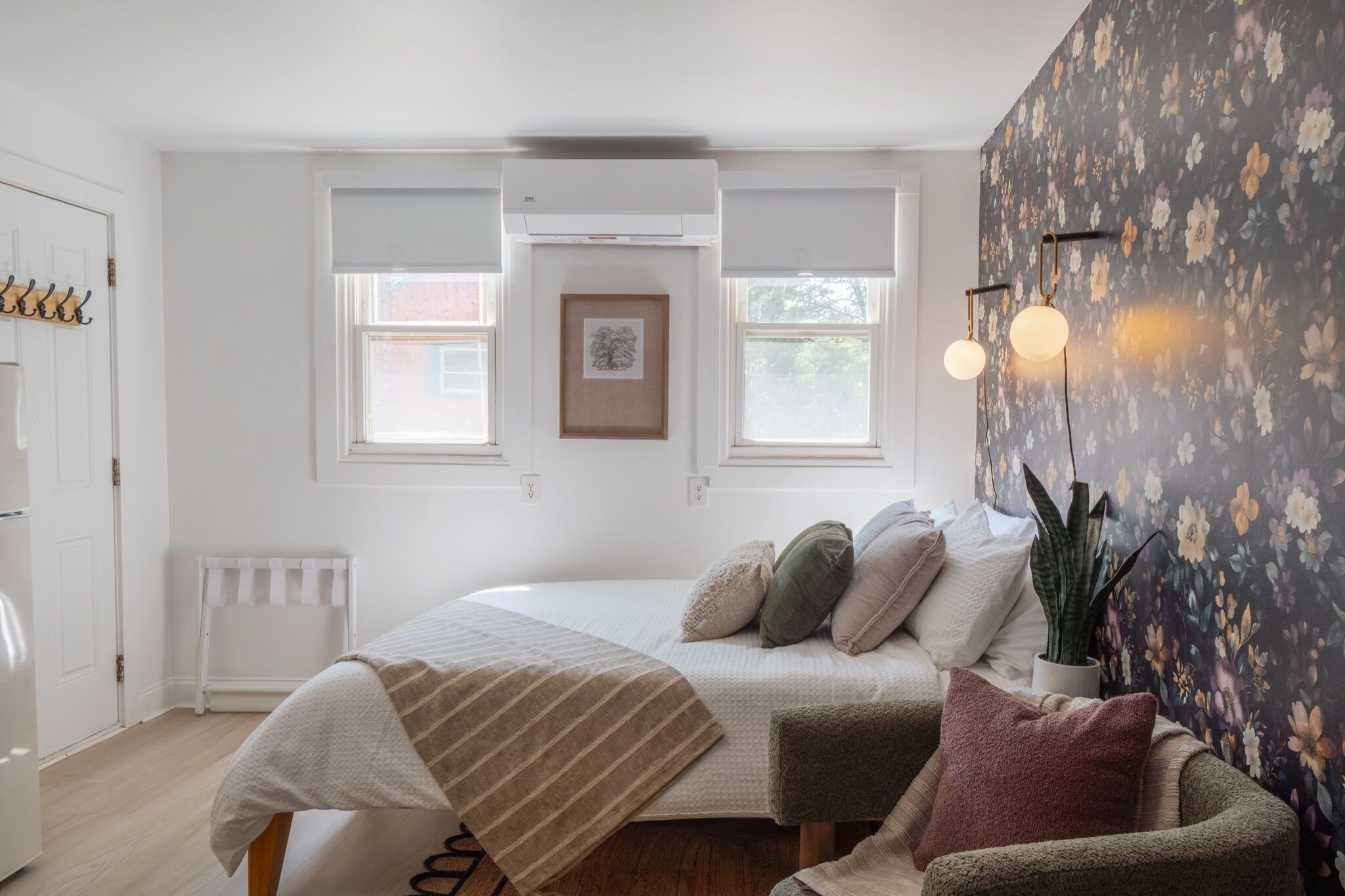 Un dormitorio con cama, almohadas, un radiador, una ventana y una pared con papel tapiz floral.
