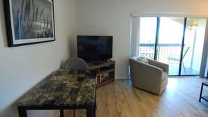 Uma sala de estar com televisão de tela plana em um suporte de madeira, um sofá, uma mesa pequena e uma varanda com vista.