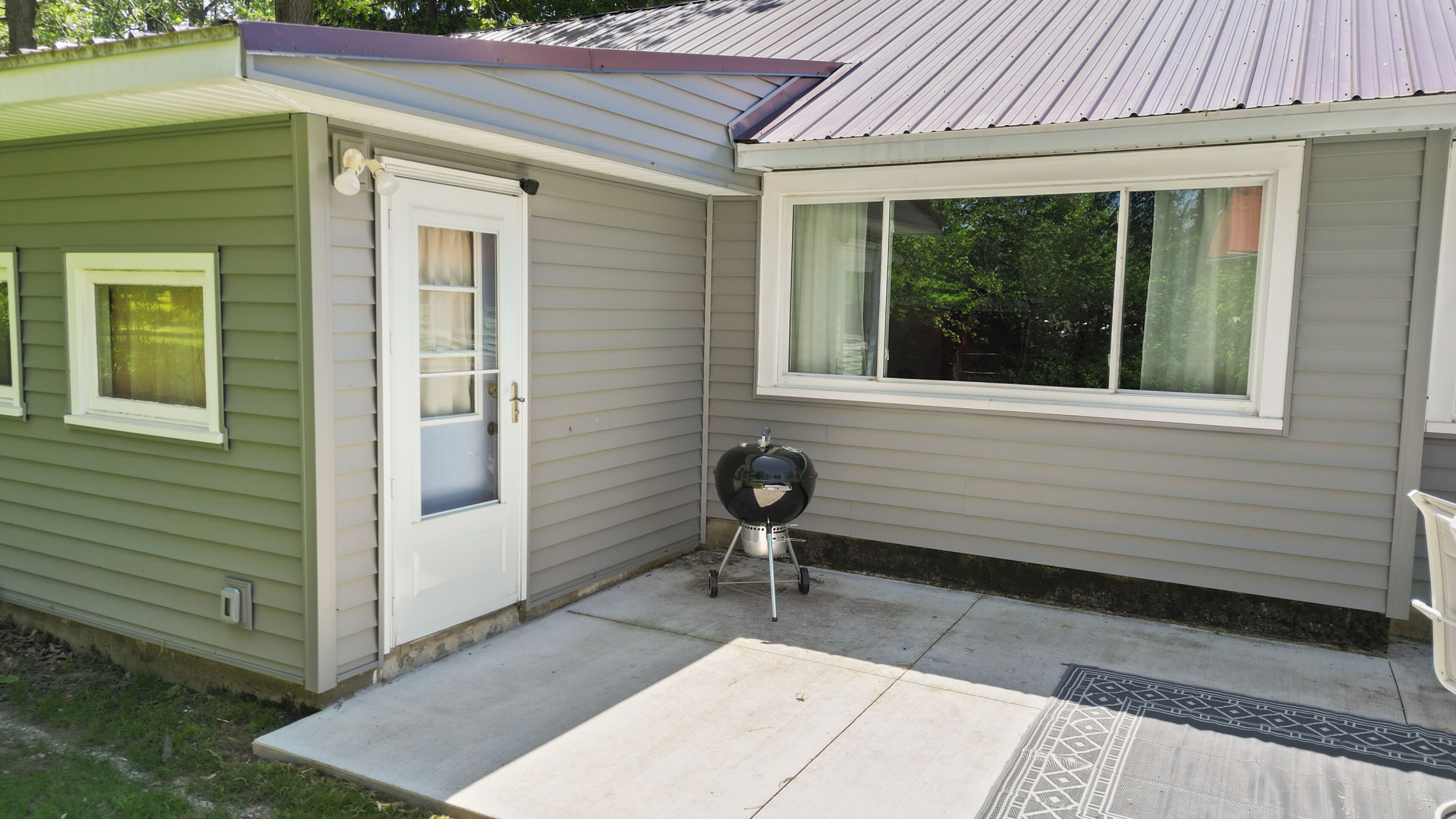 A small, single-story mobile home with a front door, a window, and a barbecue grill in the yard.