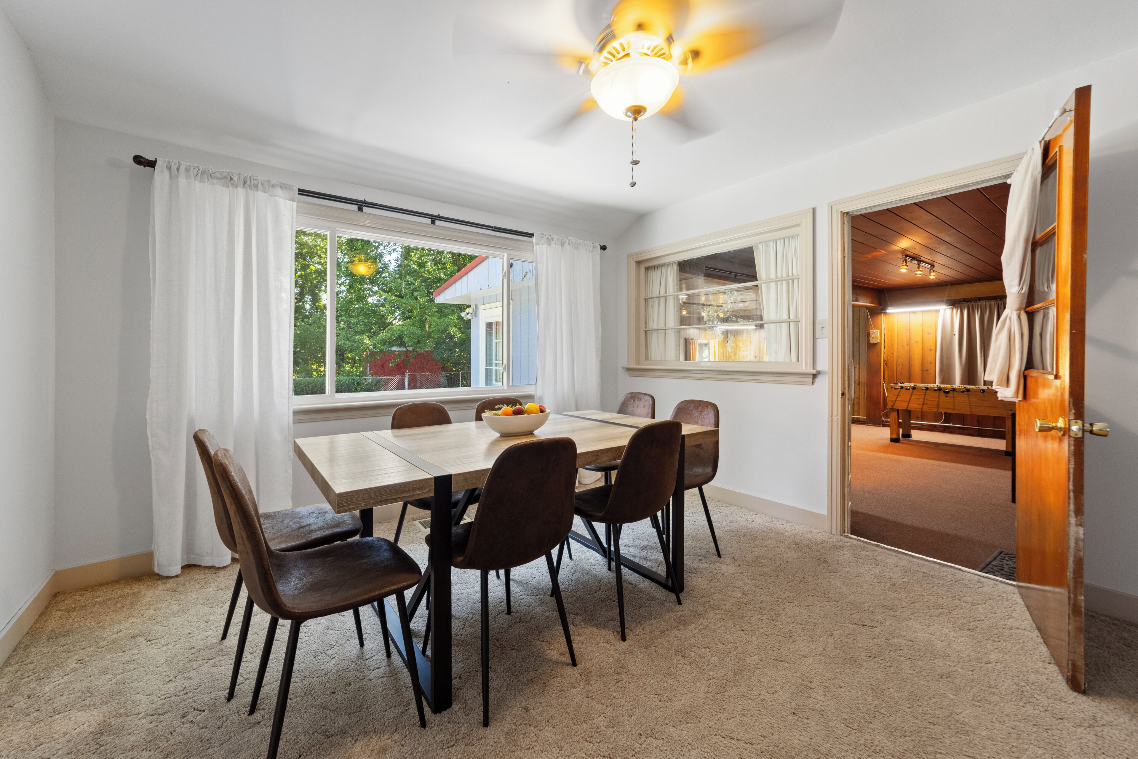 A dining area with a wooden table, six chairs, a bowl of fruit, and a view of the outdoors through large windows.