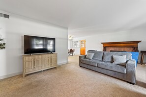 A living room with a flat-screen TV on a wooden cabinet, a grey sofa with cushions, and a wooden fireplace. - Spacious 3BR Home with Pool Table & Patio Near Lake Erie (Westfield)
