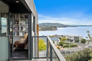 Uma varanda moderna com uma estante, uma poltrona de couro e vista para o mar e colinas.